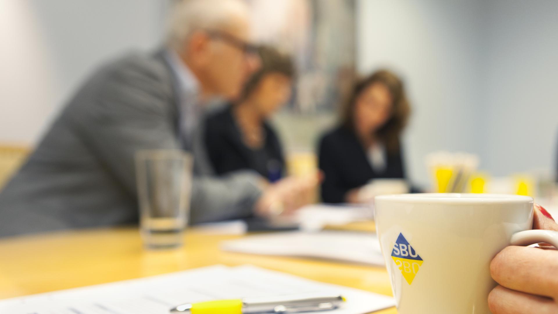 Person håller i kaffekopp i förgrunden. Personer diskuterar vid mötesbord i bakgrunden. Granskningsmall ligger på bordet