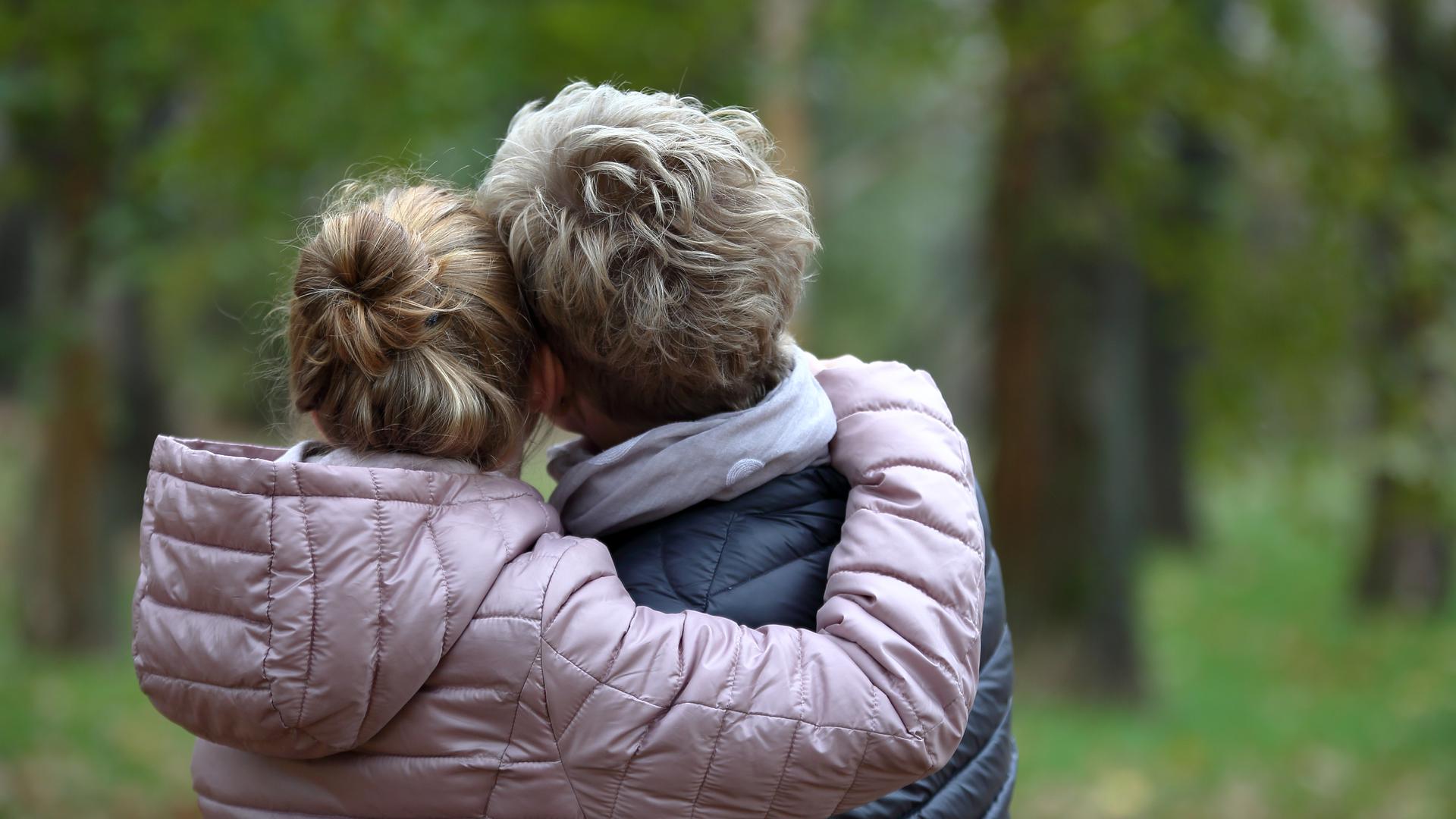 Tonårsdotter kramar sin mamma i en park. Båda står med ryggen åt kameran.