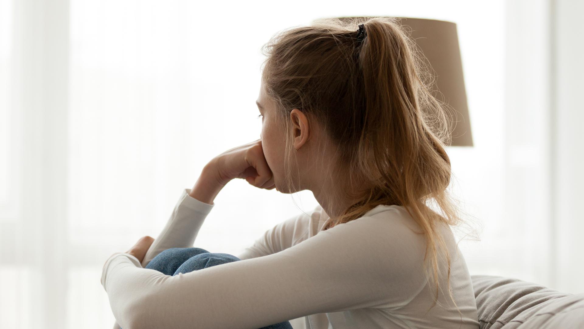 Ung kvinna sitter ensam på soffan hemma och känner sig olycklig. Hon tittar bort och ut mot fönstret.