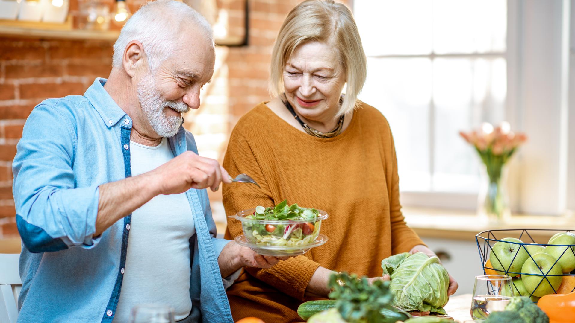 Gladlynt senior par äter sallad står tillsammans med hälsosam mat på köket hemma. Begreppet hälsosam näring i äldre ålder