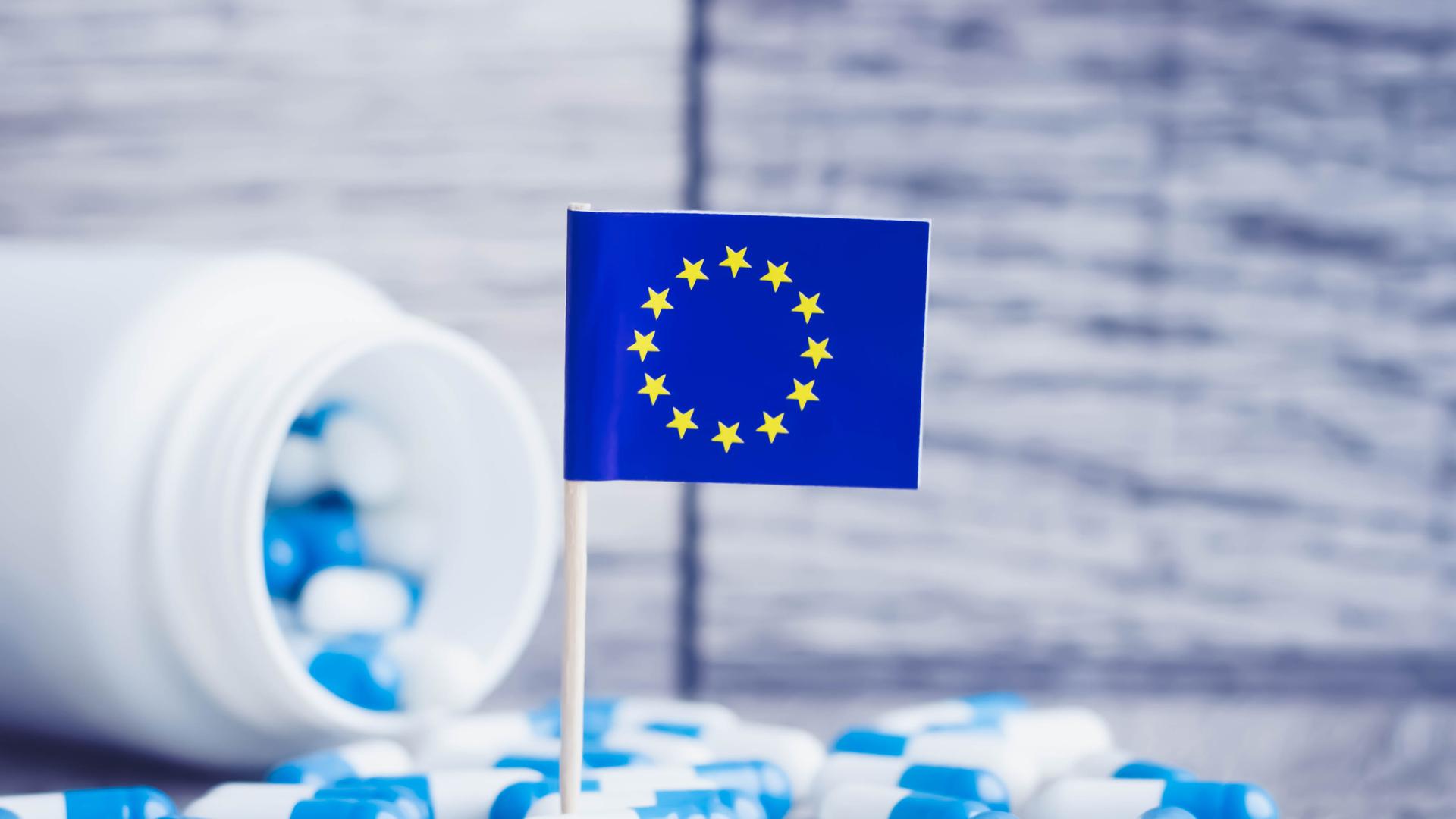 En medicinburk som vält och det ligger utspridda medicinkapslar på bordet. En EU-flagga står i mitten.