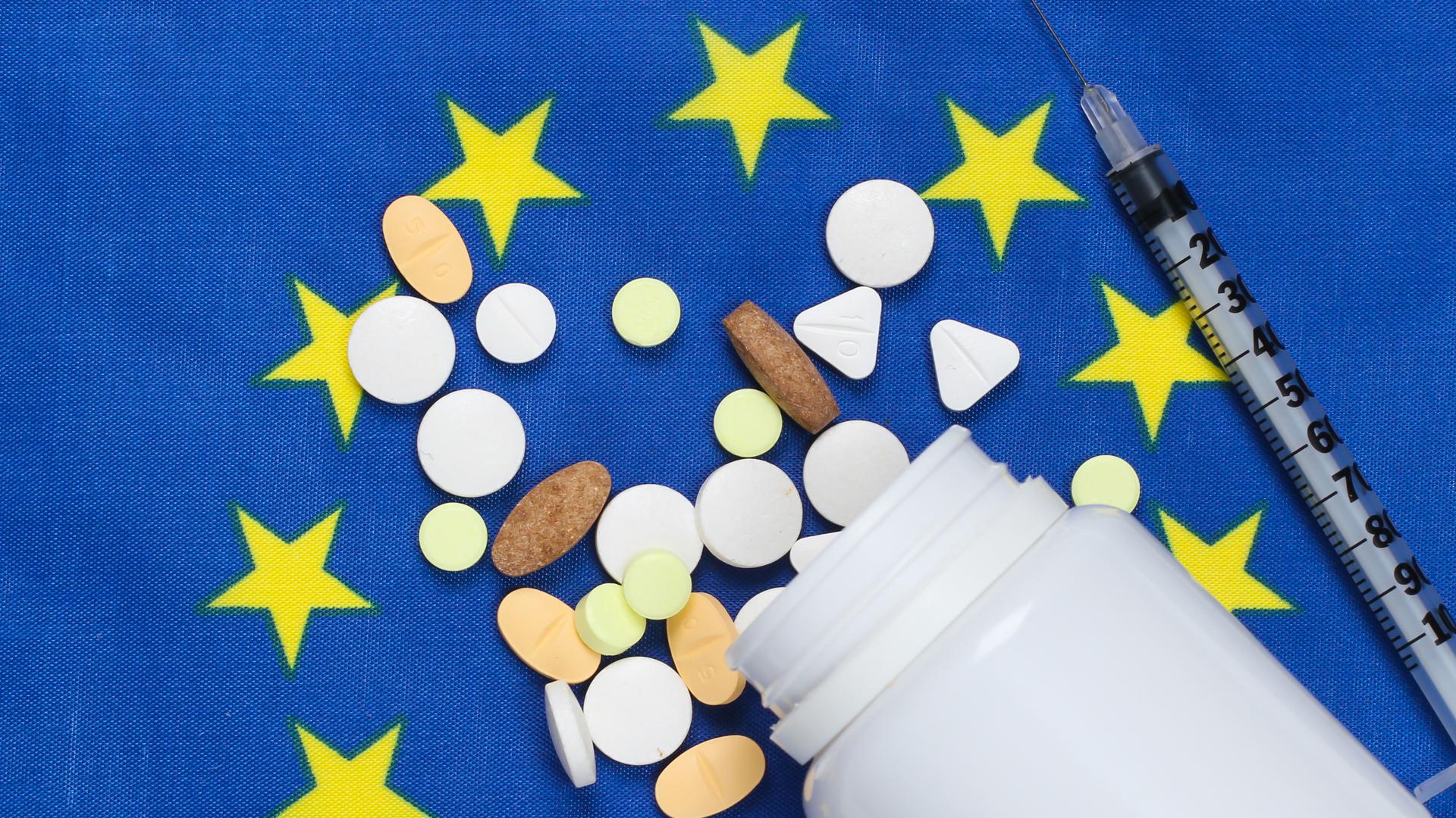 En medicinburk, olika tabletter och en spruta som ligger på en EU-flagga.