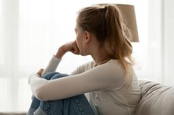 Ung kvinna sitter ensam på soffan hemma och känner sig olycklig. Hon tittar bort och ut mot fönstret.