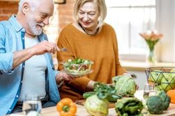 Gladlynt senior par äter sallad står tillsammans med hälsosam mat på köket hemma. Begreppet hälsosam näring i äldre ålder