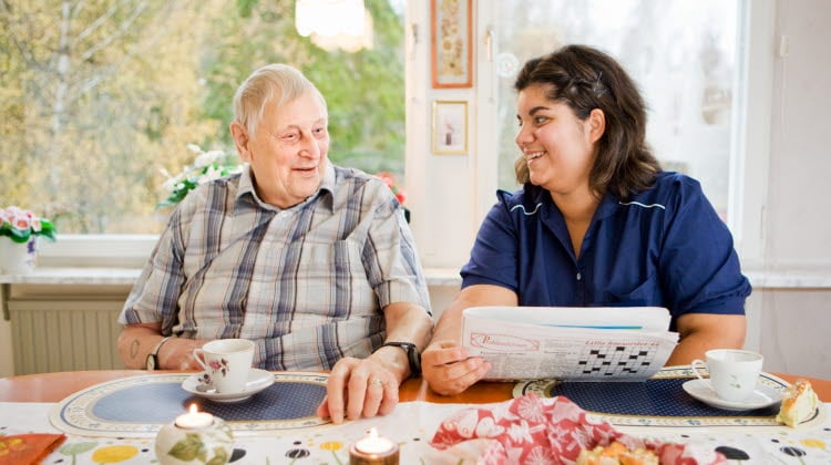 En äldre man och en kvinna ur omsorgspersonal fikar framför en tidning med ett korsord vid ett köksbord vid ett fönster med dagsljus