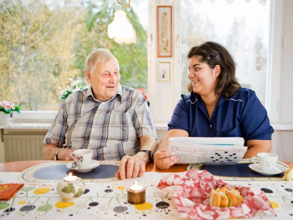 En äldre man och en kvinna ur omsorgspersonal fikar framför en tidning med ett korsord vid ett köksbord vid ett fönster med dagsljus