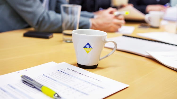 Personer vid mötesbord med vattenglas, kaffekopp och granskningsmall med penna