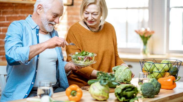 Gladlynt senior par äter sallad står tillsammans med hälsosam mat på köket hemma. Begreppet hälsosam näring i äldre ålder