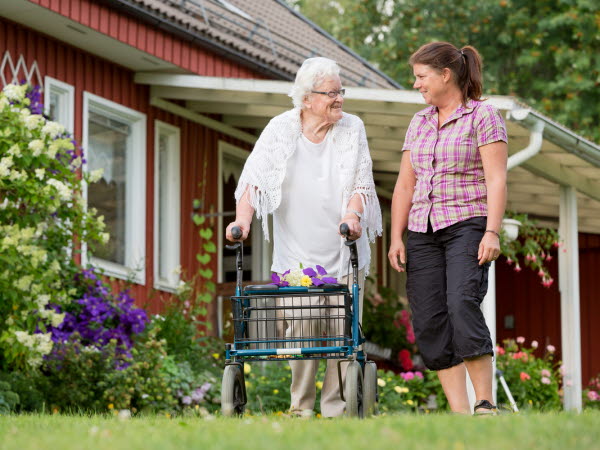 Äldre kvinna som går i trädgården med rullator och pratar med en anhörig/vårdare
