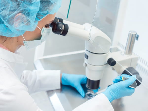 En person med hår- och munskydd, laboratorierock och handskar arbetar med en pipett och provrör under ett mikroskop.