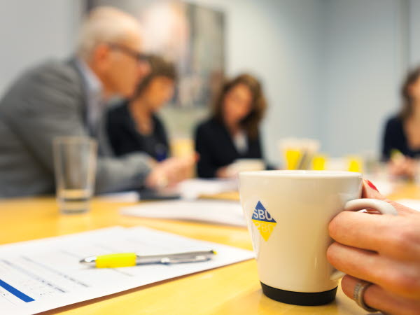 Person håller i kaffekopp i förgrunden. Personer diskuterar vid mötesbord i bakgrunden. Granskningsmall ligger på bordet